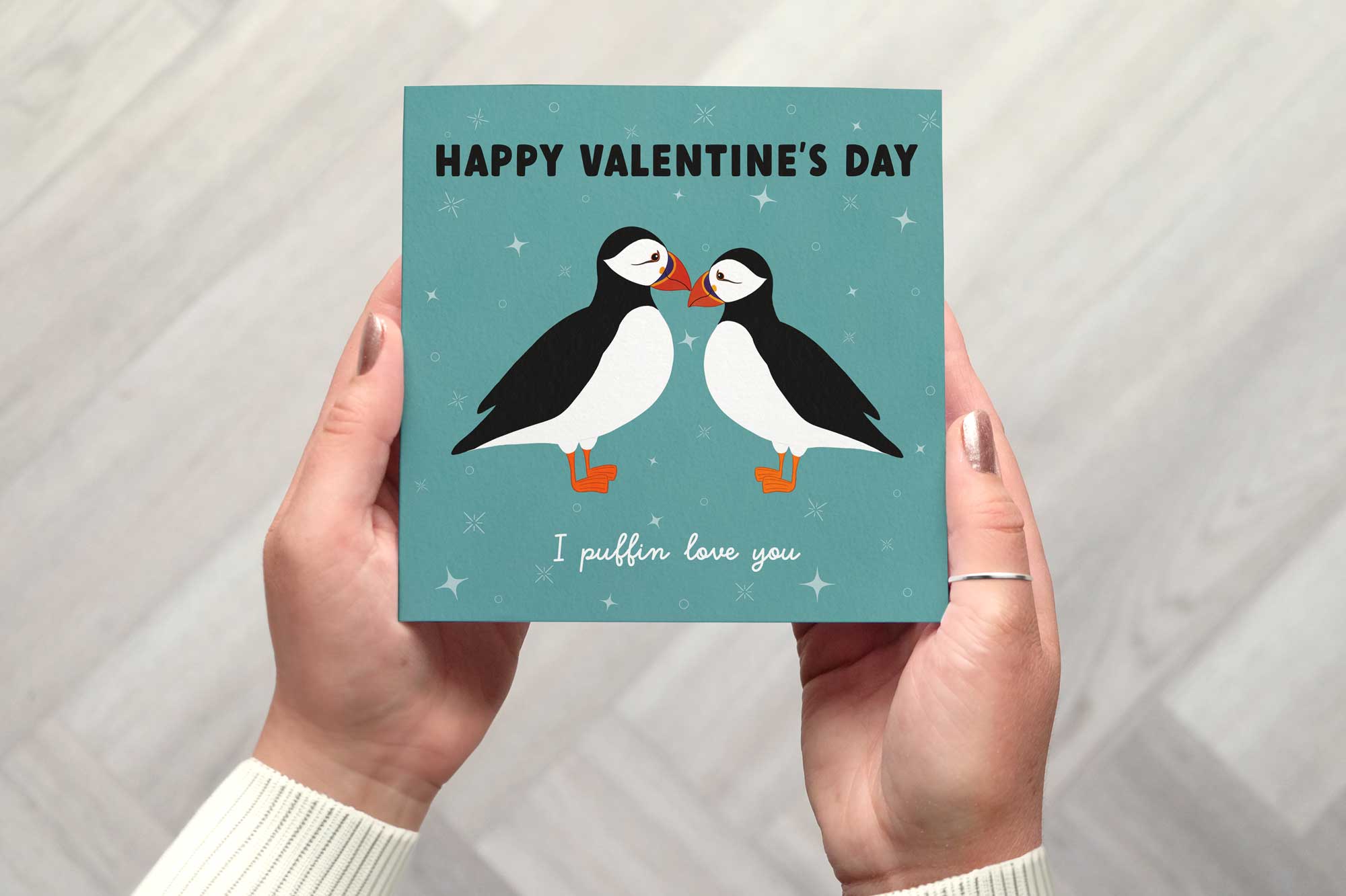 Valentines card with two puffins standing close together looking at each other. The card has a teal background with a pattern of four point stars, star bursts and circle outline. The card reads happy valentines day I puffin love you.