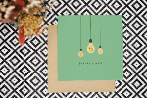 Thank you card with green background features 3 illustrated lightbulbs with cute smiling faces. The card reads Thanks a watt.