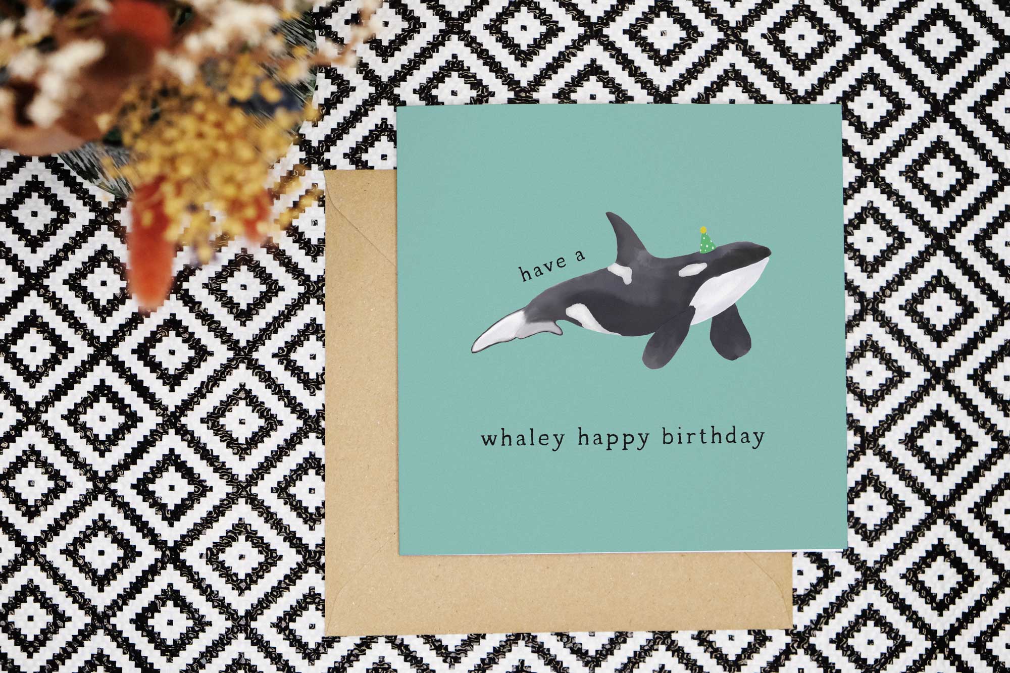 Orca birthday card featuring a watercolour killer whale wearing a green party hat. Text reads have a whaley happy birthday. The card has a teal background.