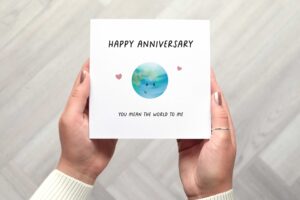 This cute anniversary card features an illustrated smiling planet earth, two pink love hearts and black text in all caps that reads Happy Anniversary you mean the world to me