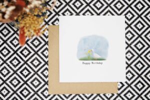 This spring themed birthday card features a white duck standing next to daffodils with two bees flying over them. It has a watercolour background of blue with green grass at the foreground. The card reads Happy Birthday under the illustration.