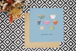 This pun 70th card has a light blue background and features seven illustrated cups of tea, each of a different type, colour and pattern. They all have cute smiling faces. The card reads seven tea.