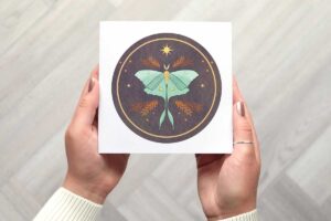 This moth card featuring a beautiful circular illustration with a dark background. it features a lunar moth with stars and moons on its wings, surrounded by stars, dots and botanical leaves.