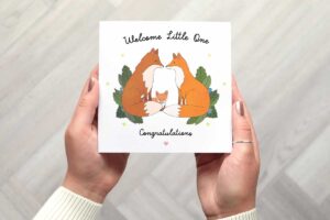 Cute new baby card with an illustration of a fox family with mum and dad sitting facing each other and baby fox seen cuddled in between them. There are leaves, flowers and stars either side of each fox. Card reads Welcome little one congratulations