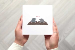This cute valentines day card features two illustrated moles looking at each other as they pop out of their mole hills with three small pink love hearts above their heads.