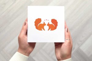 This cute valentines day card features two illustrated red squirrels sitting looking at each other with three small pink love hearts above their heads.
