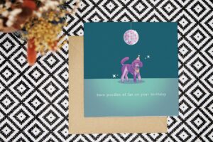 Birthday Card features a purple and lilac parti poodle illustration. The poodle wears a pink and yellow striped bandana and party hat with a yellow pom pom. Above the dog is a disco ball reflecting pastel shades of pinks, purples, blues and oranges. The card has a dark teal background with a dance floor in a lighter shade. There are some stars sparkling around the disco ball and poodle. The card reads have poodles of fun on your birthday.