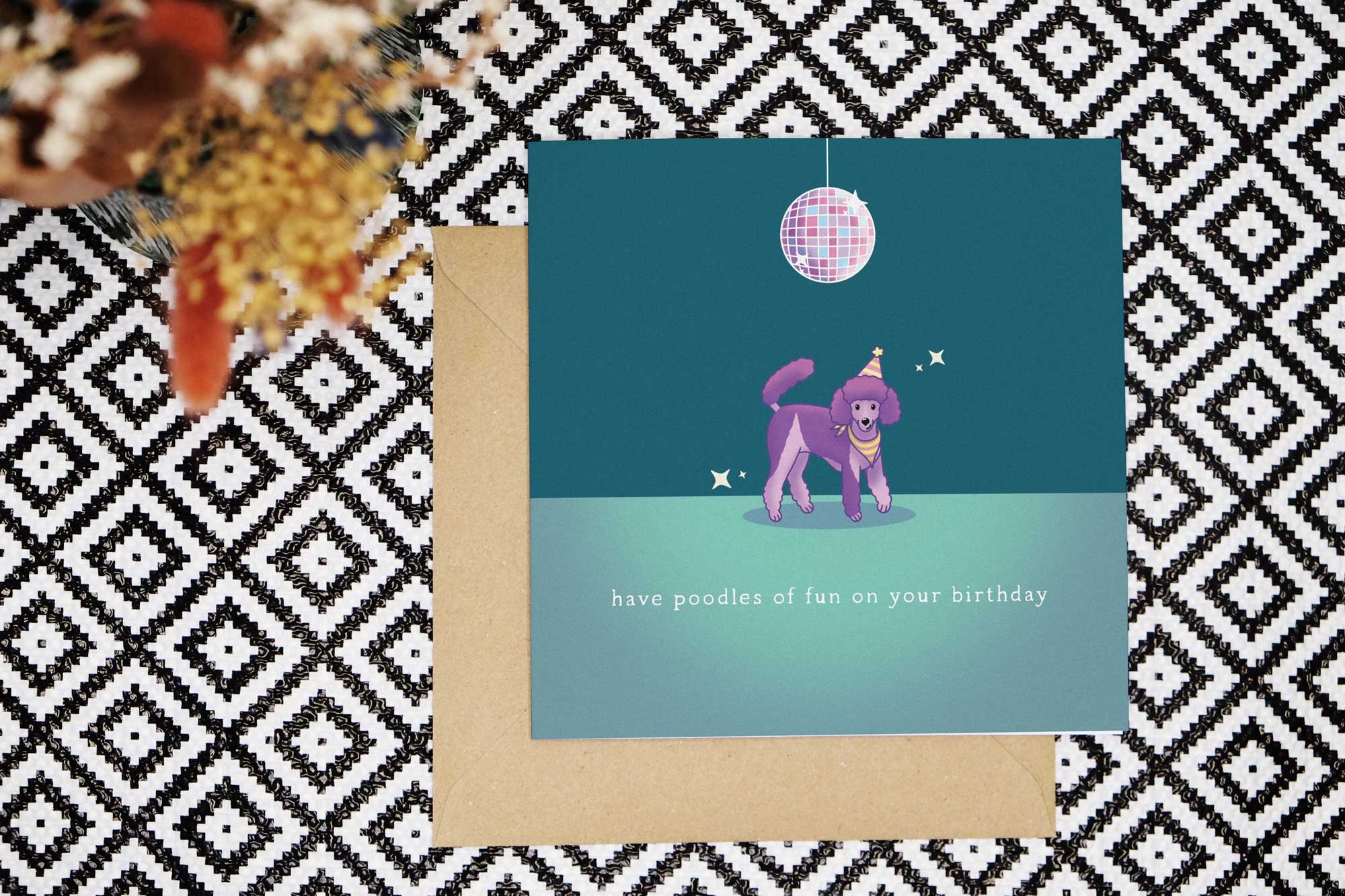 Birthday Card features a purple and lilac parti poodle illustration. The poodle wears a pink and yellow striped bandana and party hat with a yellow pom pom. Above the dog is a disco ball reflecting pastel shades of pinks, purples, blues and oranges. The card has a dark teal background with a dance floor in a lighter shade. There are some stars sparkling around the disco ball and poodle. The card reads have poodles of fun on your birthday.