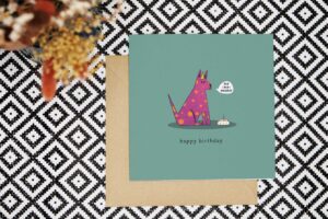 A quirky and colourful dog birthday card featuring an illustration of a purple dog with orange spots wearing an orange and yellow stripes. The dog is sitting by a dog bowl with a cake inside and a single candle. A speech bubble in front of the dog reads Hip Hip Hooray. The background of the card is teal. Underneath the illustration reads happy birthday .