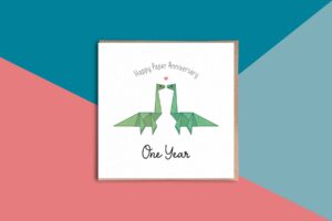 A super cute first wedding anniversary card that reads Happy Paper Anniversary One Year. Between these words are two origami diplodocus dinosaurs with a love heart above them. The dinosaurs are different shades of green. The card has a white background.