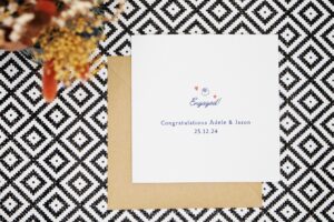 A minimal engagement card featuring an illustrated engagement ring with two love hearts either side. The card reads engaged congratulations and is personalised with the couples name and date of engagement