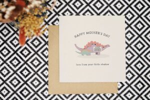 This cute mother's day card with pale yellow background features a purple mummy and green child stegosaurus. The child is following right beside its mummy saying mummy mummy mummy in a speech bubble. It reads Happy Mothers Day love from your little shadow.