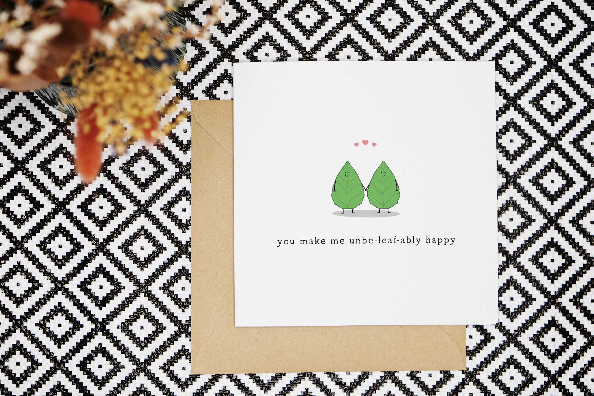 This cute pun love card features two illustrated smiling leaves holding hands with love pink hearts above their heads and the words you make me unbe-leaf-ably happy