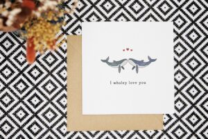 The pun whale card stood next to a succulent and vase with dried flowers. The background is botanical wallpaper and the foreground a black and white geometric pattern.