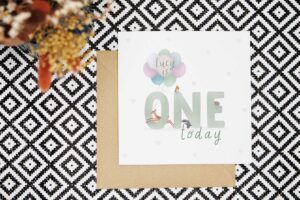 A beautiful personalised first birthday card featuring an illustration of the word one with woodland animals surrounding the letters. There is a hedgehog, badger, robin and a deer jumping through the O carrying balloons. There is a white background with light grey hearts. The card reads name is ONE today in a green text. The personalised name appears on top of the balloons. A girls name is shown in this example.
