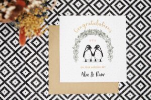 A beautiful personalised wedding card with two illustrated penguins stood under an arch of foliage. The card reads congratulations on your wedding day and is personalised with the couples names