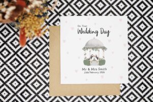 A beautiful illustrated and personalised wedding card. Featuring a hand illustrated gazebo decorated with garlands of pink, purple and white flowers, foliage and hedges. There are two puffins stood in the centre. The card reads on your wedding day and is personalised with the couples name and date of the wedding. The card has a white background with faint pink love hearts.