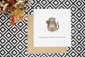 A cute get well soon card featuring an illustrated tabby cat holding a bouquet of sunflowers with a red ribbon in its mouth. The pun hope you are feline better soon is written underneath the cat illustration.