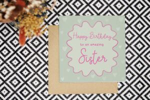 A green birthday card that has a border of hand drawn pink squiggle around the text with a lighter green inside to make the text pop. It reads happy birthday to an amazing sister. The text is pink. There are pale green love hearts featured in the darker green background of the card.
