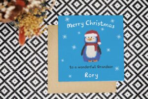 This personalised Christmas card features a hand drawn penguin wearing ared striped scarf and a red Santa hat with white trim and pom pom. Created using oil pastels and embellished with hand drawn digital background and snowflakes. The card reads Merry Christmas to a wonderful and can be personalised with the relation and name