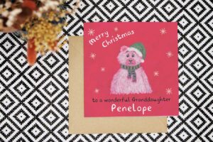 This personalised Christmas card features a hand drawn red background and polar bear wearing a green striped scarf and a green Santa hat with white trim and pom pom. Created using oil pastels and embellished with hand drawn digital snowflakes. The card reads Merry Christmas to a wonderful and can be personalised with the relation and name