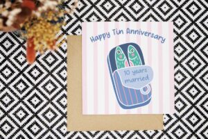 Celebrate 10 years married with this colourful and playful card with an illustrated tin of sardines. There are two sardines inside and the tin has been opened to review the words 10 years married on the inside of the lid. The card has a pink and white striped background and the illustration features bright blues, pinks and greens. The card reads Happy Tin Anniversary.